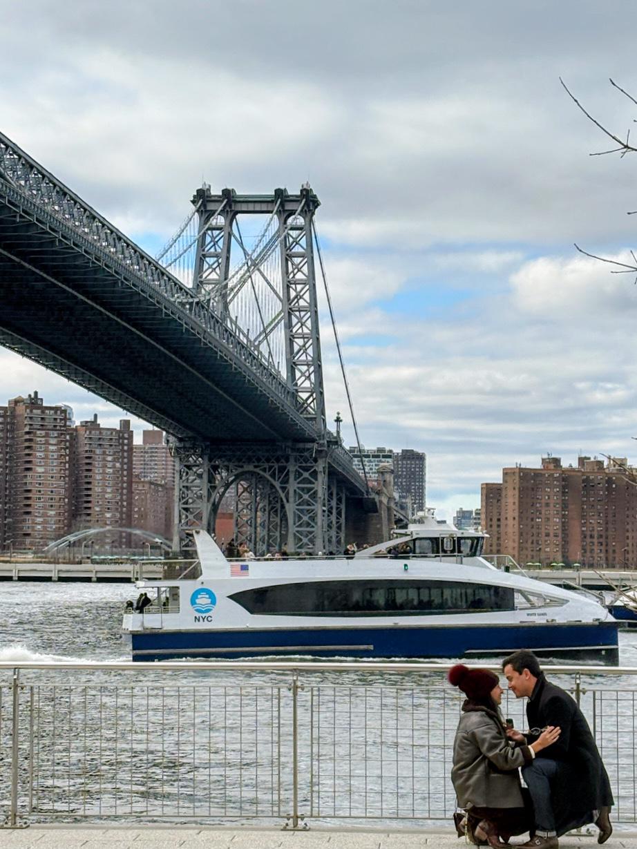 Alexei proposing to Ximena at Domino Park in Brooklyn with the Williamsburg Bridge and East River behind them